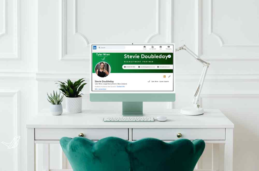 A picture of a green scallop-edge chair sitting empty in front of a white desk and white-panelled wall. On the desk sit 2 small house plants in white pots, 1 angle-poise white desk lamp, and a screen showing a perfected LinkedIn profile