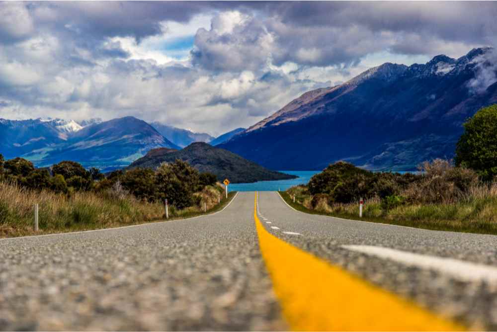 Photograph from a camera sitting on a paved road on New Zealand's South Island. The road (with a bold yellow central marking) leads down to a blue lake, surrounded by green hills and snowy moutains in the distance beneath a cloudy sky, representing the Public Practicce Accounting Opportunities available on the South Island.