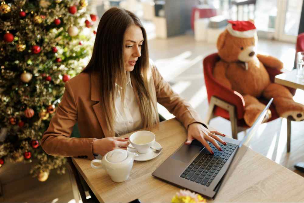 Photograph of a woman with long brown hair in a white shirt working on a laptop on a desk, infornt of a Christmas tree. She is holding a cup of coffee and next to her in a chair is a teddy bear wearing a Christmas hat, showing a year-end job search